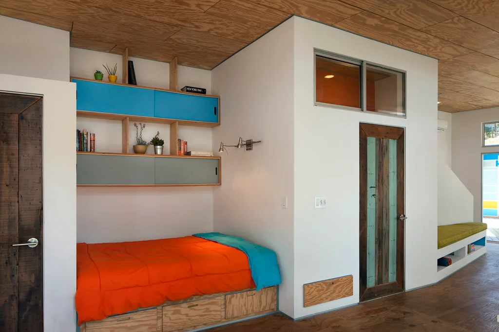 Built-in bed nook with an orange cover under blue shelving and plywood ceiling