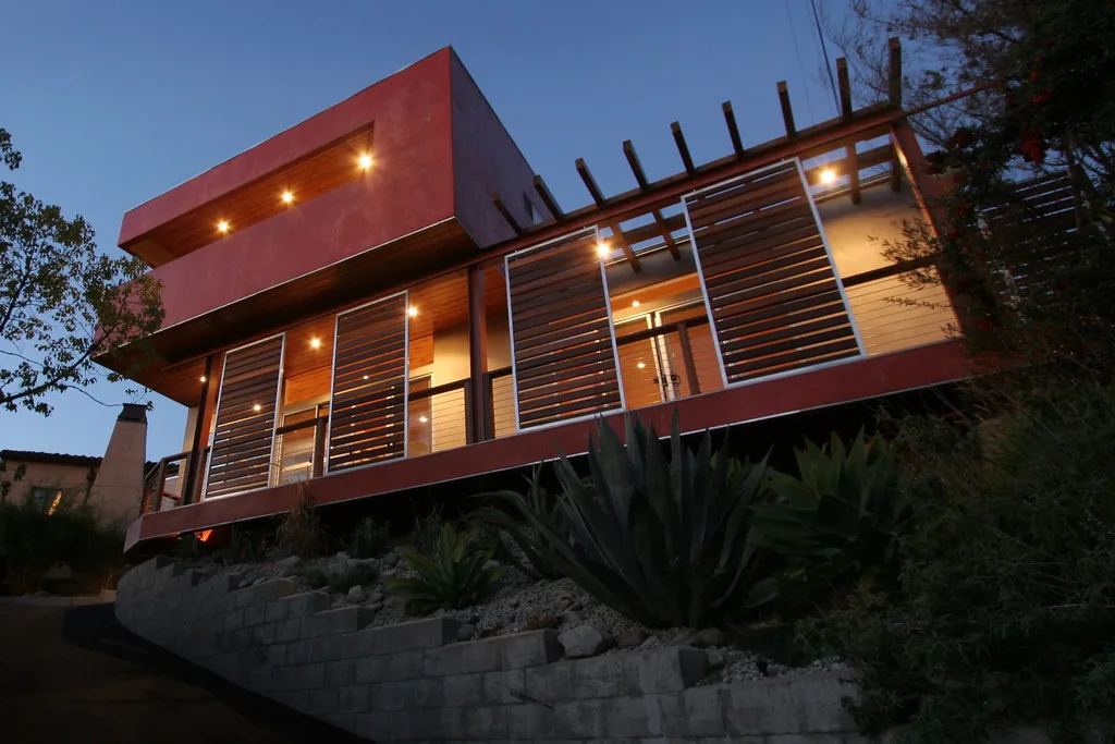 Modern house with a red cantilevered upper volume and wood sunscreens lit at dusk