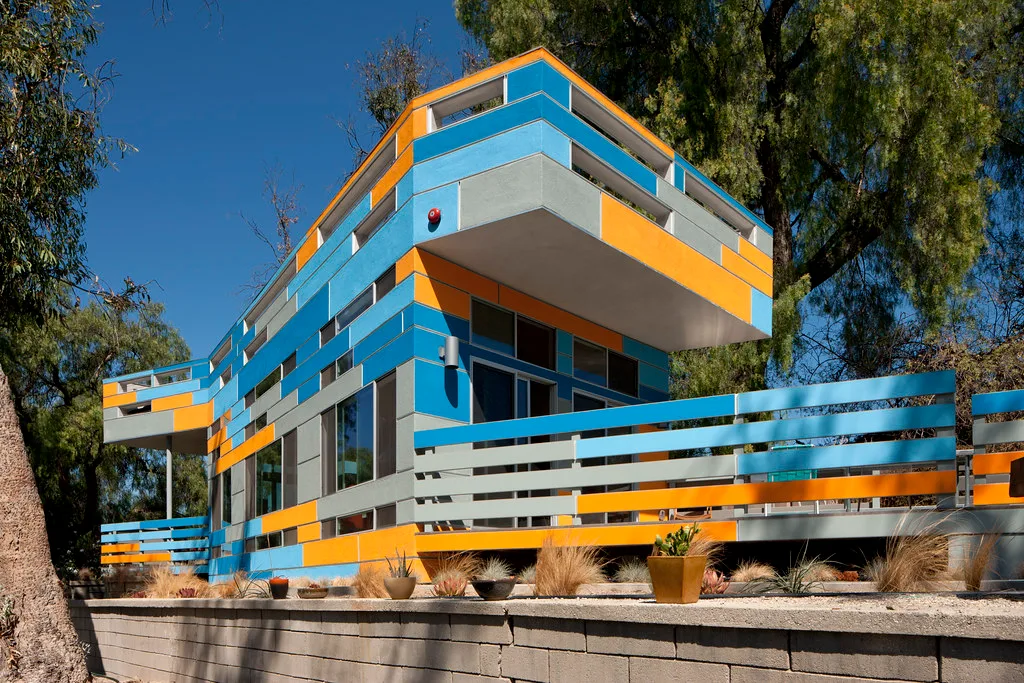 Modern hillside house clad in blue, orange and grey panels with a cantilevered upper floor