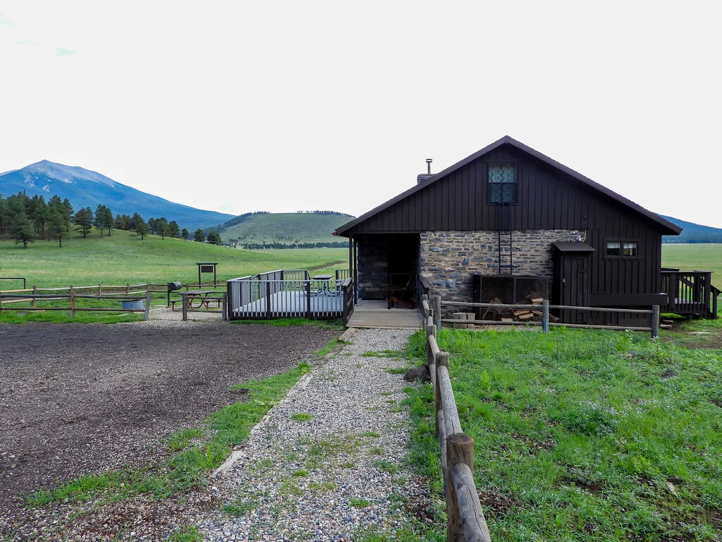 Stone and dark-timber cabin with a deck, set in a green meadow below a mountain peak