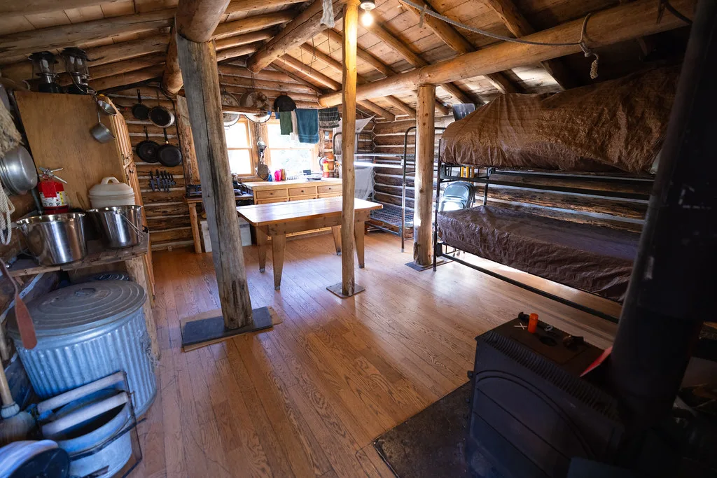 Long log room with a wood stove, plank table and bunks under timber beams